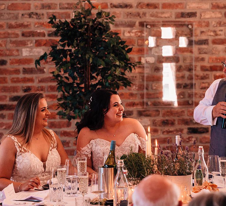 The brides beam brightly at the father of the bride performing his wedding speech 