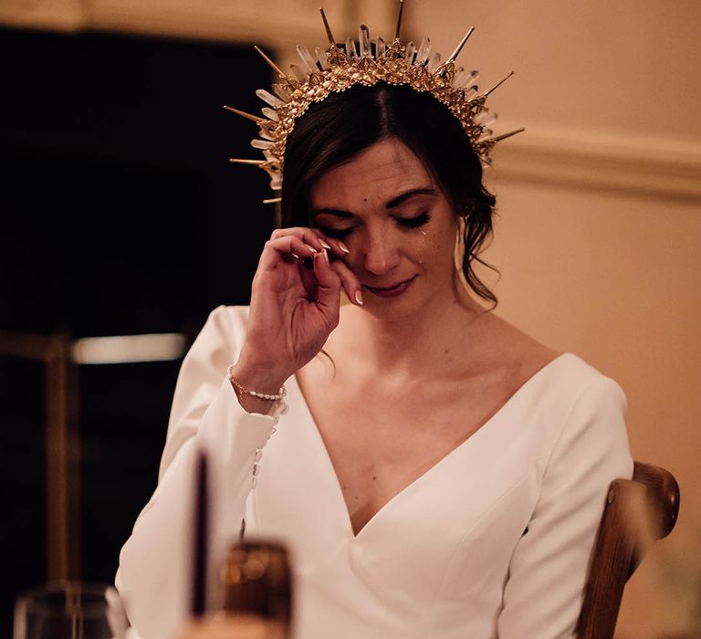 Bride in long sleeve wedding dress wipes tears during the wedding ceremony as she feels emotional wearing a gold bride crown 