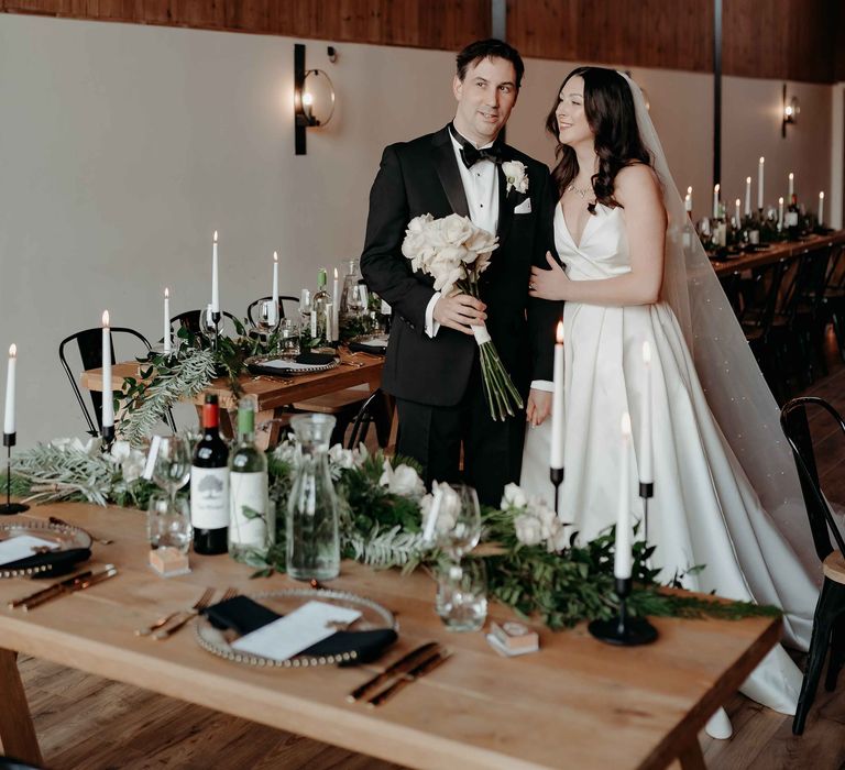 Bride in white silk wedding dress and veil stands with groom in black tuxedo at wedding breakfast featuring tablescapes of green foliage, white-tapered candles and wine bottles