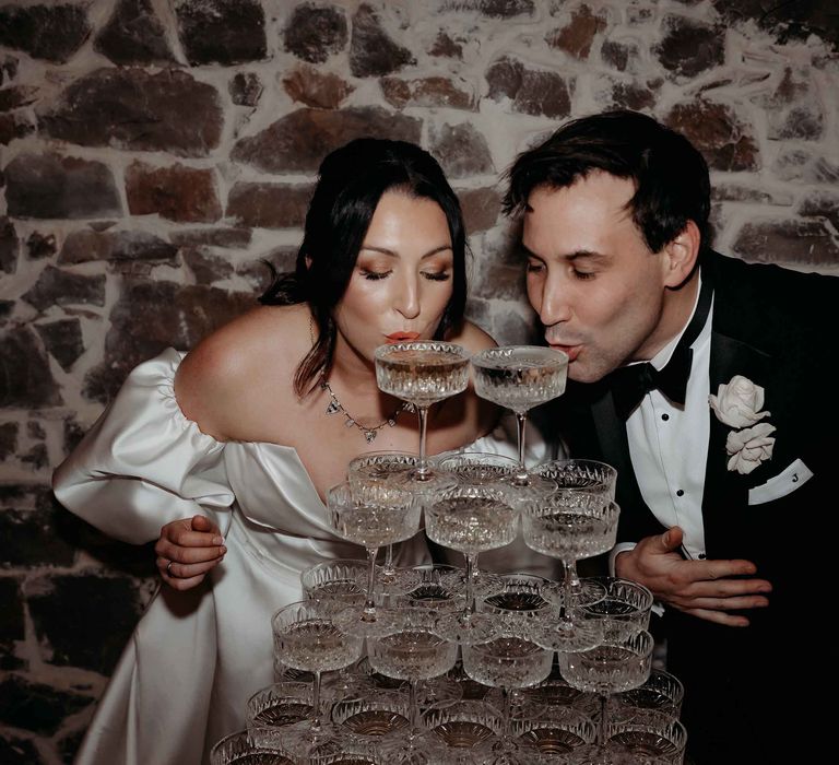 Bride in puff sleeve wedding dress and groom in black tuxedo sipping from a champagne tower at Upton Barn wedding venue