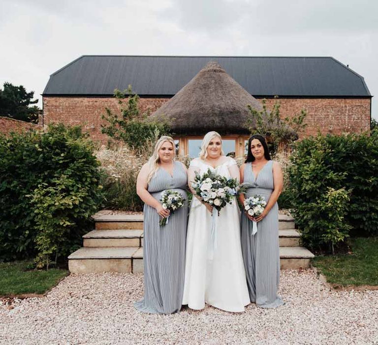 Bridesmaids wearing dusky blue sleeveless crinkle bridesmaid dresses standing with bride in satin off the shoulder wedding dress holding white and blue eucalyptus, foliage, garden rose, gardenia, light blue delphinium and dried flower bridal bouquet tied with dusky blue ribbon 