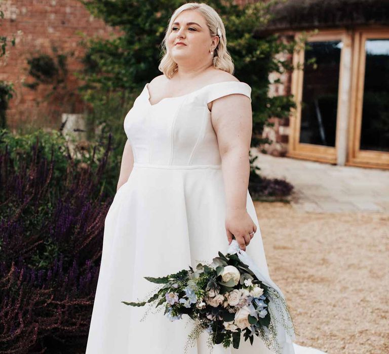 Bride walking around the grounds of Brickhouse Vineyard Devon wedding venue wearing satin off the shoulder wedding dress holding white and blue eucalyptus, foliage, garden rose, gardenia, light blue delphinium and dried flower bridal bouquet tied with dusky blue ribbon 