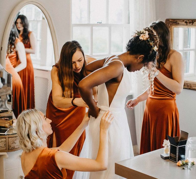 Bridesmaids in one shoulder burnt orange bridesmaid dresses helping bride into her v-neck sleeveless wedding dress with 3D floral applique train at Brinsop Court wedding