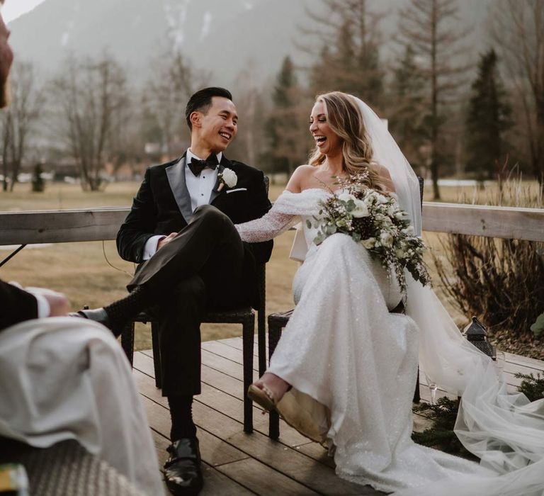 Bride in off shoulder sparkly mermaid wedding dress holding white garden rose, eucalyptus, baby's-breath and foliage bridal bouquet laughing at the alter with groom in classic black tuxedo, black bowtie and white garden rose boutonniere 