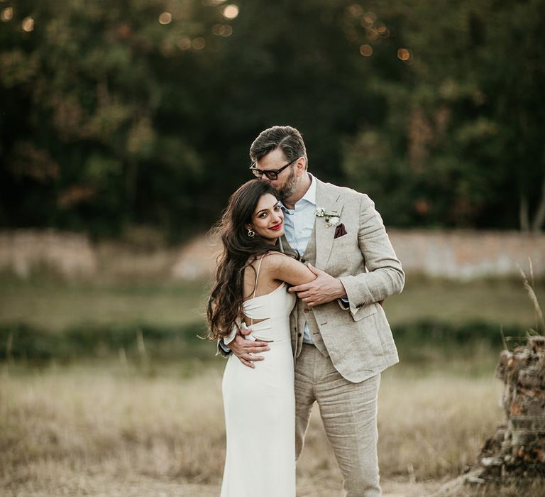 Groom in three piece cream wedding suit with navy blue tie embracing bride in fitted slip wedding dress with tousled brown hair 