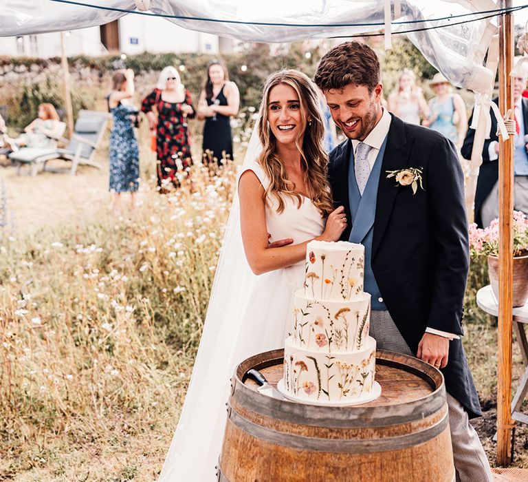 bride and groom cutting the three-tier flower pressed wedding cake displayed on a barrel 