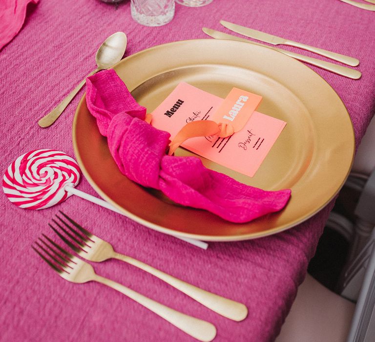 Gold charger plate with hot pink napkins, coral place name and lolly favours 