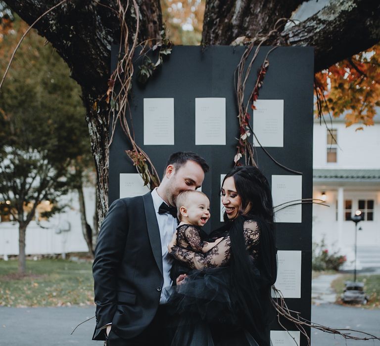 Bride in black wedding dress holding baby girl in matching black dress with the groom in front of wedding table plan 