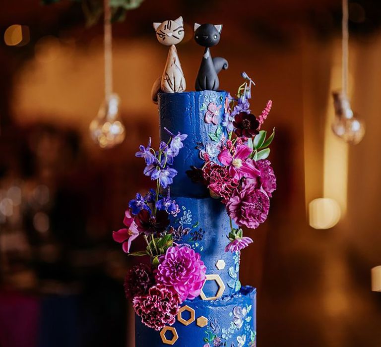 Extravagant dark blue and purple wedding cake with flowers, cat cake toppers and gold hexagonal detailing 