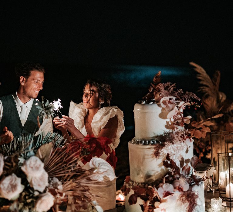 Outdoor wedding reception on the beach in Italy with giant wedding cake with dried flower decor and bride and groom holding sparkles 