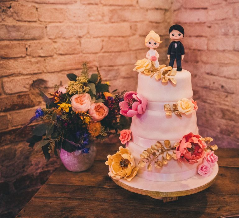 White three tiered wedding cake with pink and yellow flower decorations and bride and groom figurines