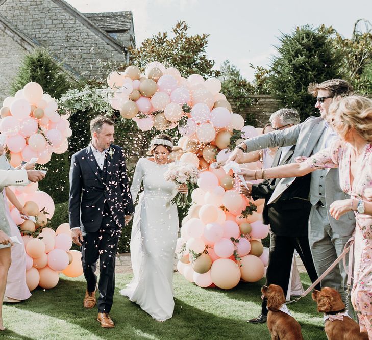 Bride in Elbeth Gillis gown and groom in navy Cad & the Dandy suit walk through guests throwing confetti in front of pink and gold balloon arch at Euridge Manor wedding