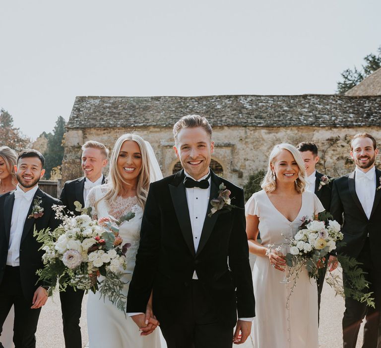 Bride in white Charle Brear wedding dress holding hands with groom in black velvet Hugo Boss suit with groomsmen and bridemaids