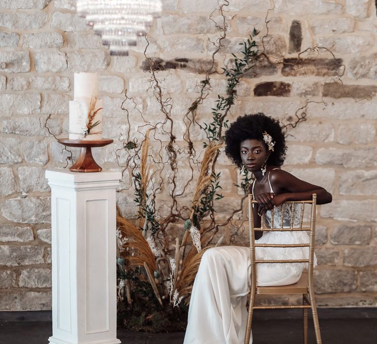 Black bride in a fitted wedding dress sitting on a chair at Ponden Mill with chandelier light installation, two-tier wedding cake on a plinth and dried flower and foliage floral arrangement 