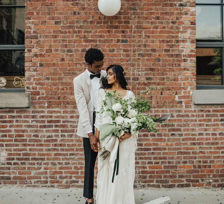 Bride and groom portrait by a brick wall with the boho bride holding an oversized white and green wedding bouquet 