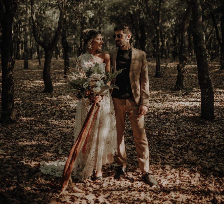 Groom in a beige suit and black t-shirt standing with his boho bride in a lace one shoulder wedding dress in a forest 