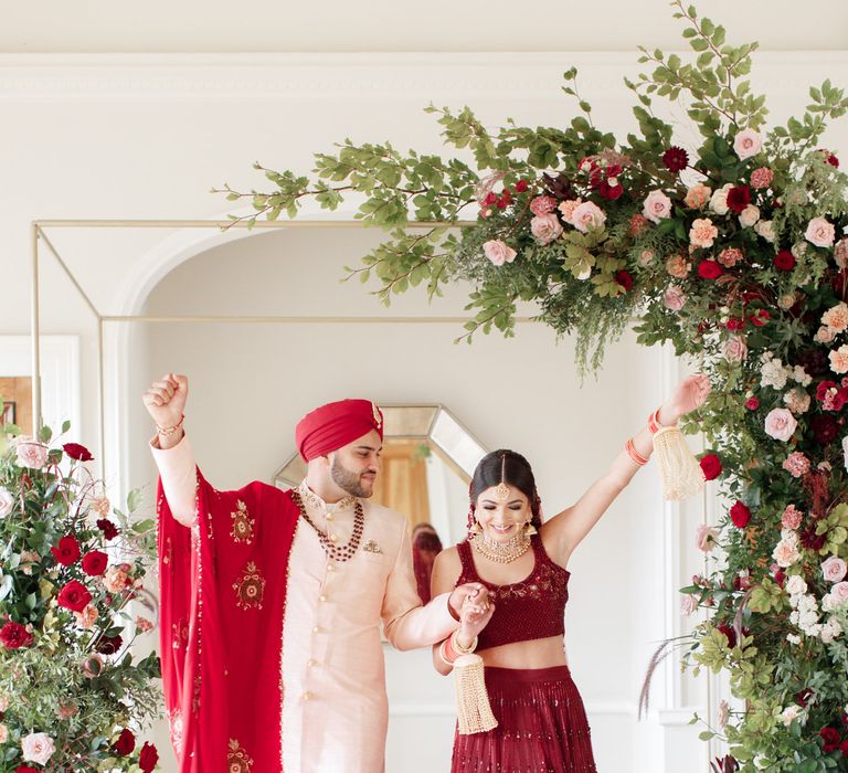 Sikh couple celebrate under floral archway filled with roses and green foliage