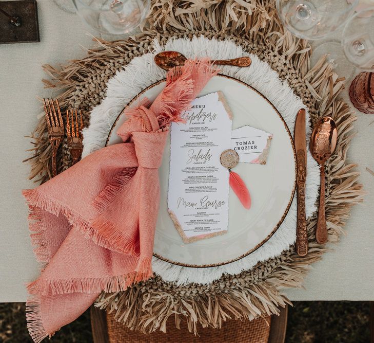 Place setting with wicker place mat, gold cutlery, coral napkin and menu card