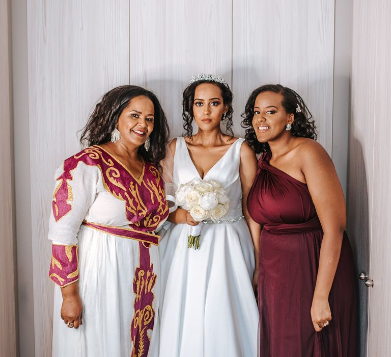 Bride in a princess wedding dress and tiara holding a white peony bouquet standing with her mum and sister in a burgundy dress