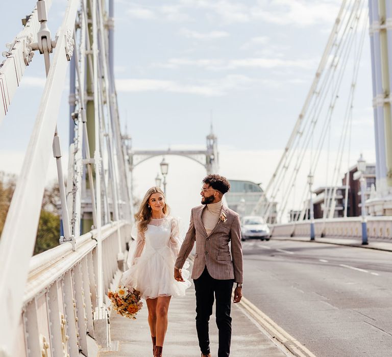 Bride in a short ruffle wedding dress with boots walking across Albert's bridge with her groom in a brown check blazer 