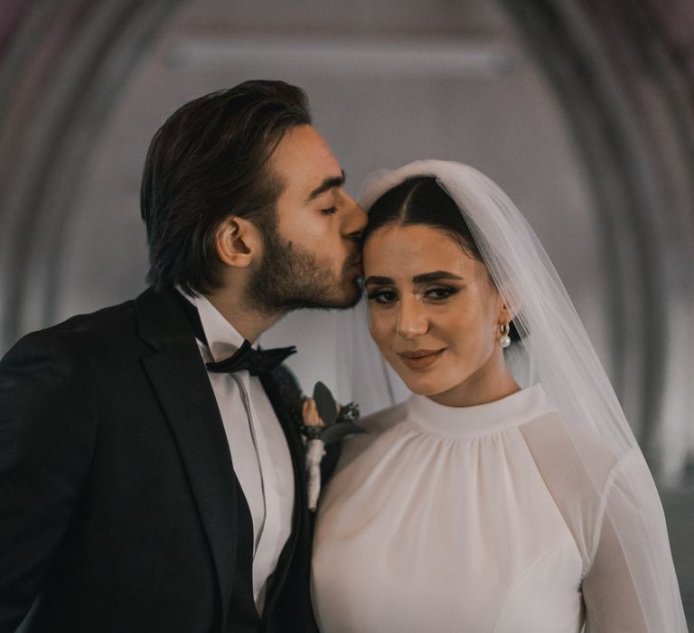 Groom kisses the cheek of his bride as she smiles at the camera 