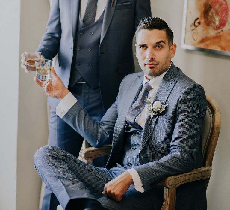 The groom and best man in matching three piece blue suits, toasting with a drink