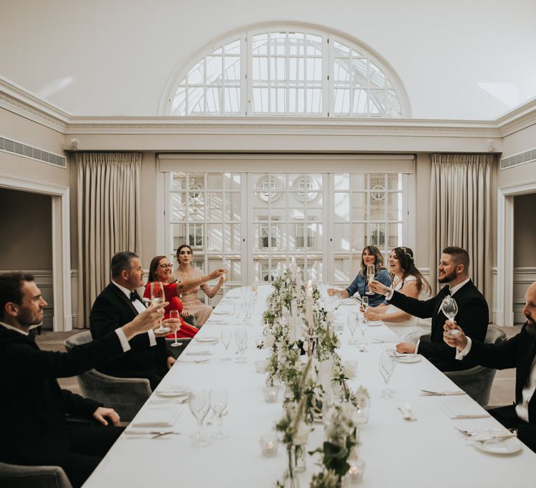 Wedding party sit around table for intimate dinner as table is decorated with a white tablecloth and small floral arrangements