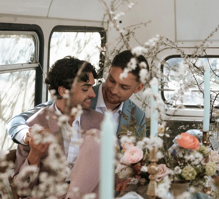 Grooms sit together surrounded by rustic floral decor and candlesticks in brass holders 