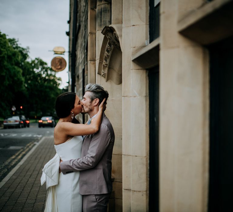 Bride in white strapless Rebecca Vallance Dress with bow detail kisses groom in light brown Moss Bros suit and blue tie on the stress of Harrogate after wedding