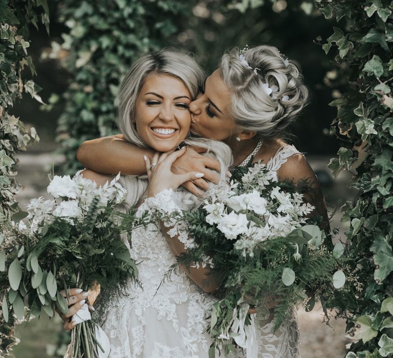 Two brides in white dresses holding white and green bouquets embrace under trees at crab & lobster fairytale wedding. 