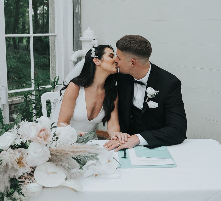 Bride with long dark hair held back with silver hair accessory wearing white Pronovias wedding dress kisses groom in black tuxedo as they sit signing the wedding registry at Came House Dorset wedding