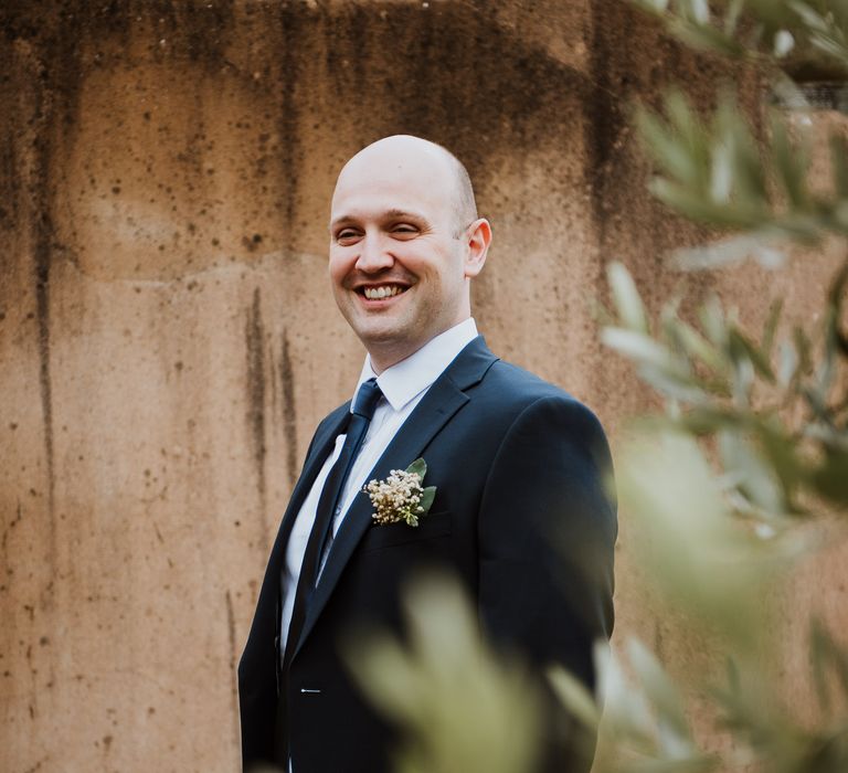 Groom stands behind florals as he wears suit with simple floral buttonhole within his pocket