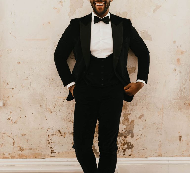 Smiling groom in a black velvet tuxedo standing against plain industrial style wall