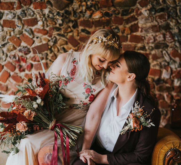 Two brides sitting on a vintage armchair, one bride is wearing a long veil cape dip dyed orange for their pumpkin spice wedding