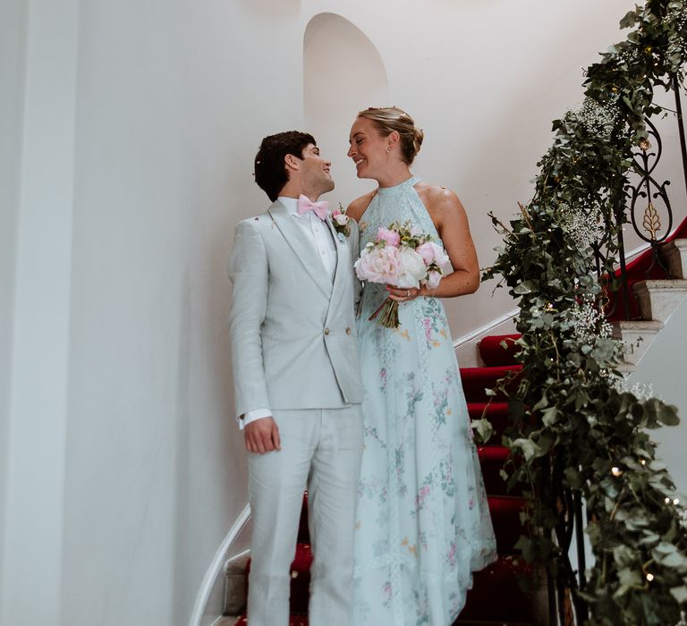 Bride & groom stand on staircase lined with green foliage and florals on the day of their wedding