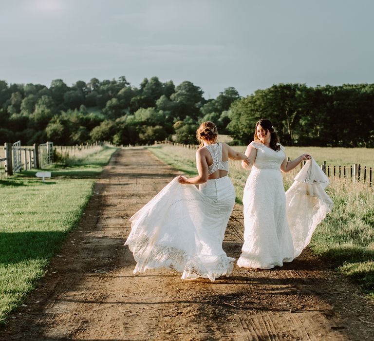 Brides walk through countryside on their wedding day as they lift their skirts and dance with one another