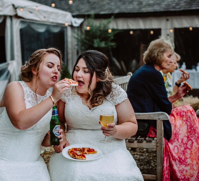 Bride feeds her bride pizza on the day of their wedding as they sit outside at Park Farm