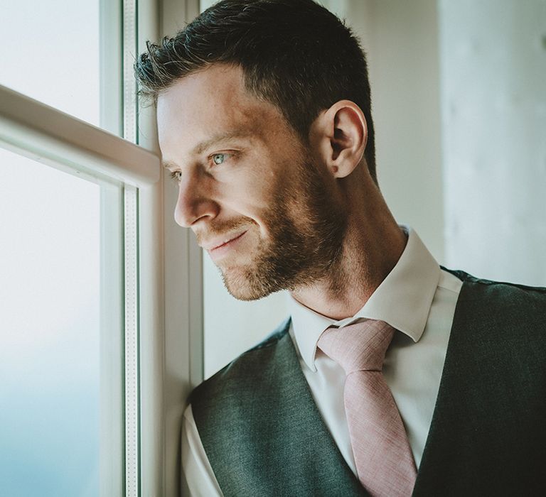 Groom looks out the window as he wears pale pink tie and grey waistcoat