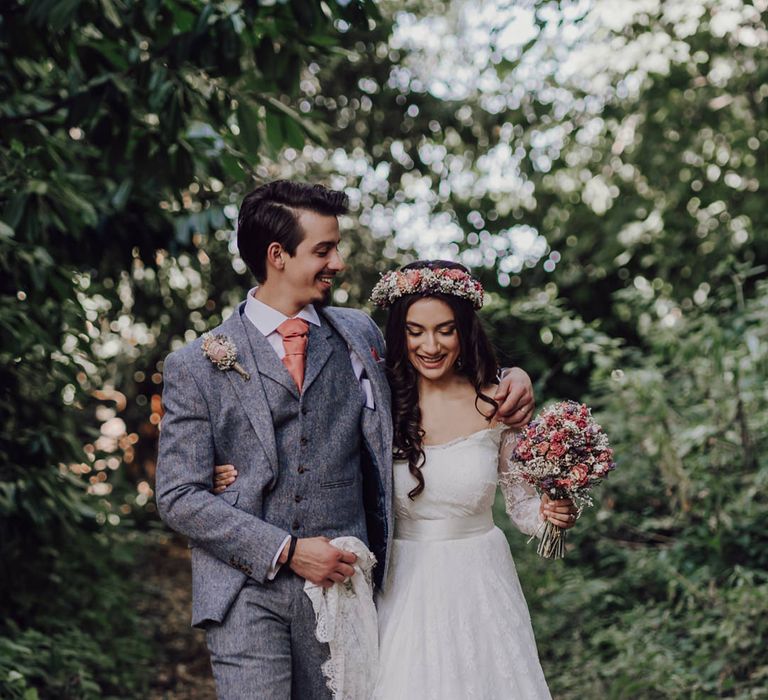 Groom in a three-piece grey wool wedding suit and coral cravat holding his brides lace wedding dress train 