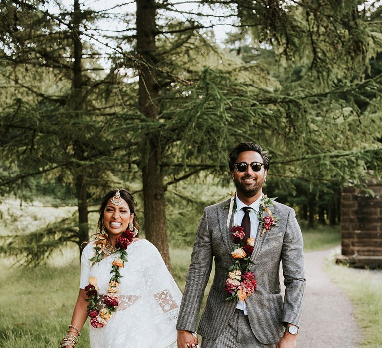 An Indian British bride and groom walk to their wedding breakfast holding hands and smiling. He wears a grey suit and she wears a white sari and carries a colourful bouquet.