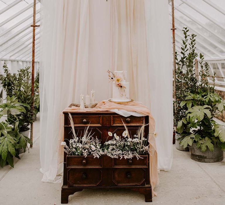 Two tier buttercream wedding cake sitting on a vintage dresser in front of a large white drape in the glasshouse at Gooseberry House 