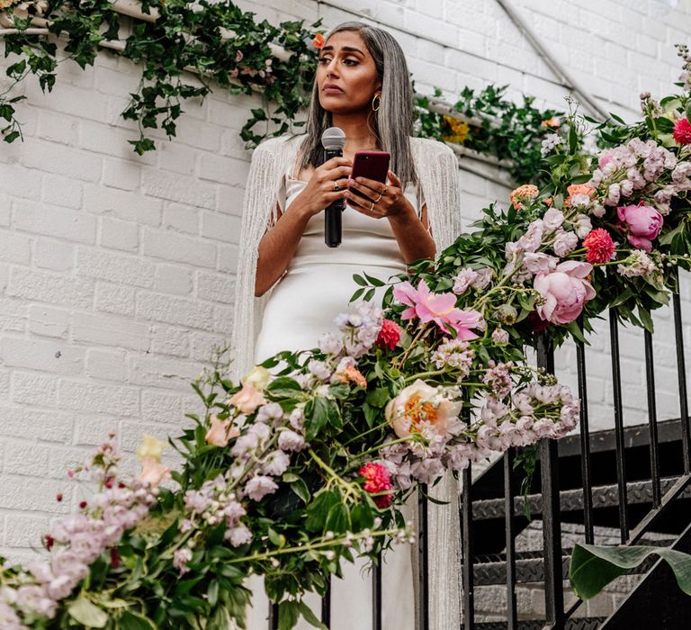 Bride in satin Halfpenny London wedding dress and tasselled bridal cape holds phone and microphone as she stands on metal stairs with floral banister decoration during wedding reception at Loft Studios London