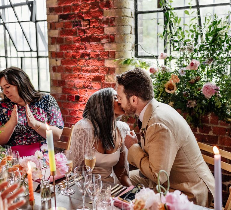 Bride in satin Halfpenny London wedding dress with tasselled bridal cape kisses groom in linen suit during wedding breakfast in room of exposed brick with candles, florals and wooden chairs