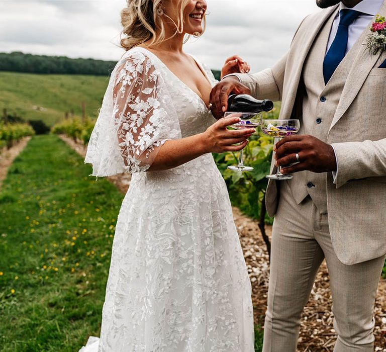 Bride in a lace wedding dress and groom in beige suit with blue check pouring champagne into couple glasses 