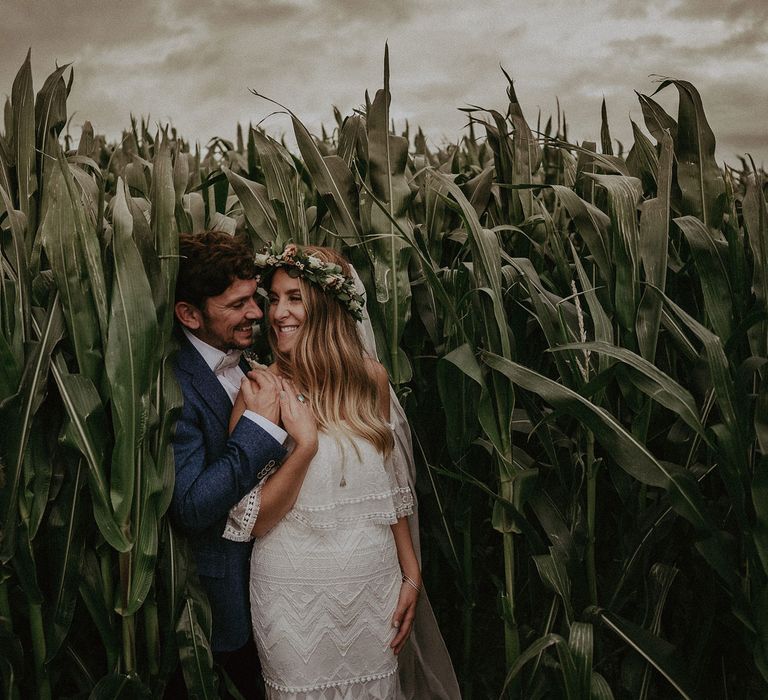 Bride in Grace Loves Lace bardot wedding dress, flower crown and veil stands with groom in blue suit jacket in corn field after Isle of Wight wedding with macrame wedding decor