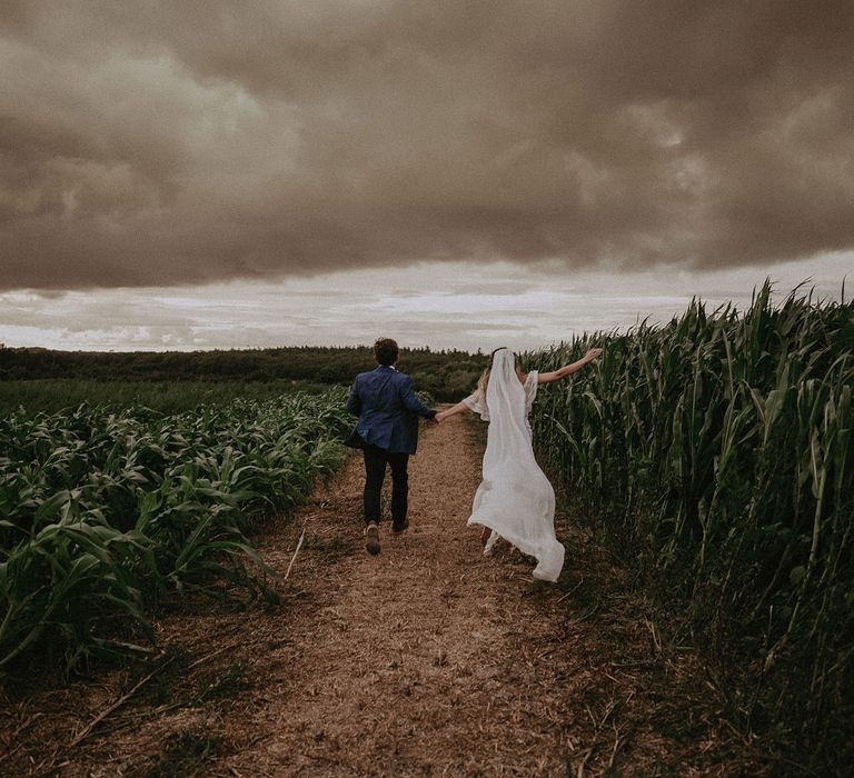 Bride in Grace Loves Lace wedding dress and veil runs through corn field holding hands with groom in blue suit jacket at Isle of Wight wedding with macrame wedding decor