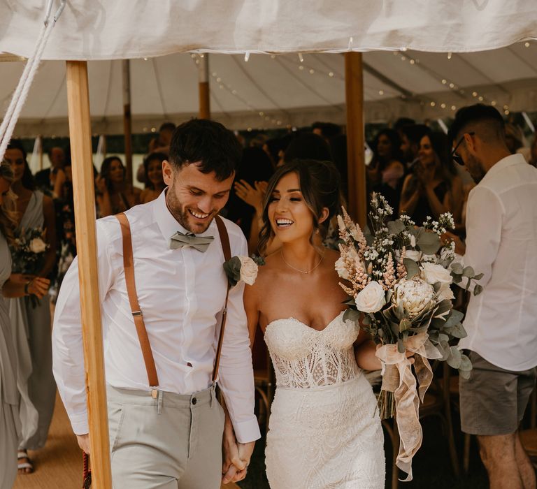 Bride & groom hold hands as they walk through marquee and bride holds her floral bouquet | Mark Bamforth Photography