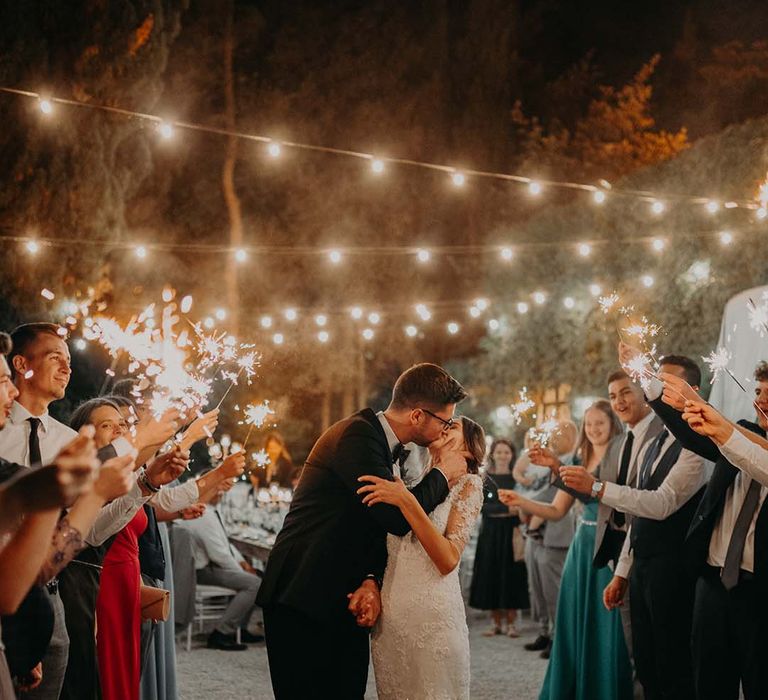 Bride & groom kiss as wedding guests hold sparklers and fairy lit canopy hangs above them | Royal Studio