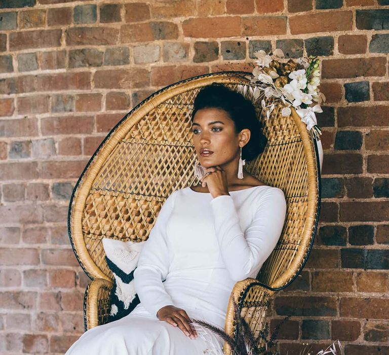 Stylish bride in a minimalist wedding dress with slash neck and long sleeves sitting in a peacock chair at a boho black tie wedding 