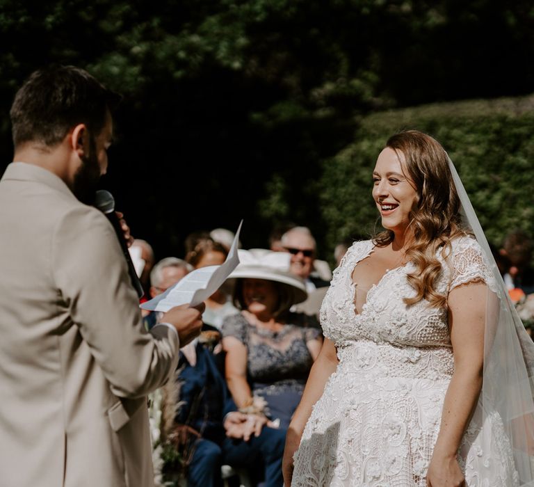 Bride in homemade lace wedding dress and veil laughs as groom in beige suit reads out vows during DIY garden wedding ceremony in Bedfordshire